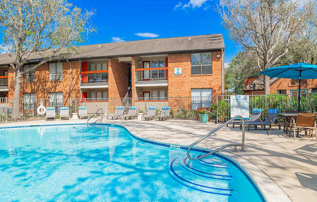 A swimming pool in front of a brick building with a blue umbrella.
