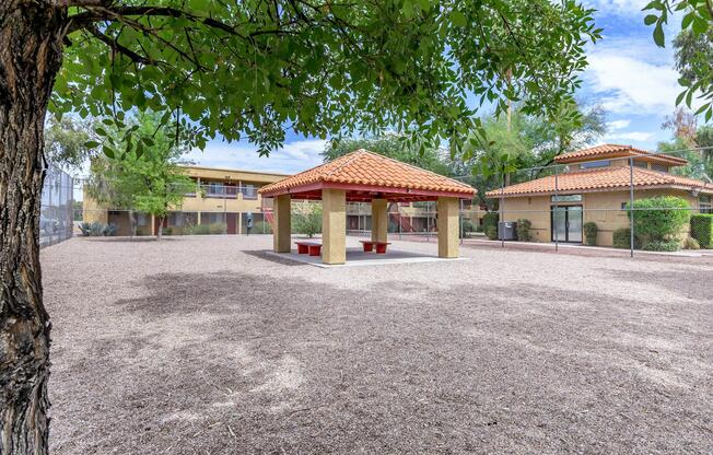 A shaded gazebo with a tiled roof sits in a gravel area surrounded by trees and buildings. The scene is bright with clear skies, showcasing a peaceful outdoor space ideal for relaxation or gatherings.