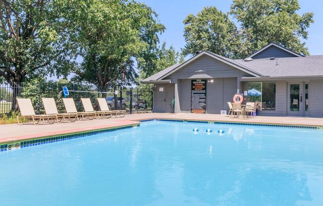 A clear blue swimming pool surrounded by lounge chairs, with a shaded area and a nearby building featuring large windows. Trees and a fence are visible in the background, and a sign can be seen on the building. The scene is bright and inviting, suggesting a recreational area for relaxation and leisure.