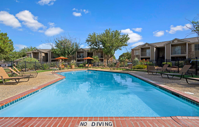 A sunny outdoor swimming pool here at Northridge Court surrounded by a spacious pool deck with lounge chairs, patio umbrellas, and landscaped greenery, with nearby apartment buildings, mature trees, and a clear blue sky creating a relaxing community setting.