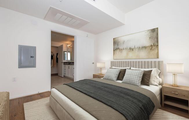 A softly lit bedroom featuring a neatly made bed with grey and beige bedding. On either side of the bed, there are wooden nightstands with lamps. A large piece of abstract art hangs on the wall above the bed. The room has a doorway leading to another area and a wall-mounted electrical panel. The decor is modern and minimalistic, with light-colored walls and warm wooden flooring.