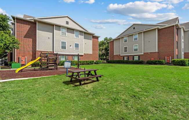 A grassy area in front of apartment buildings with a picnic table and playground.