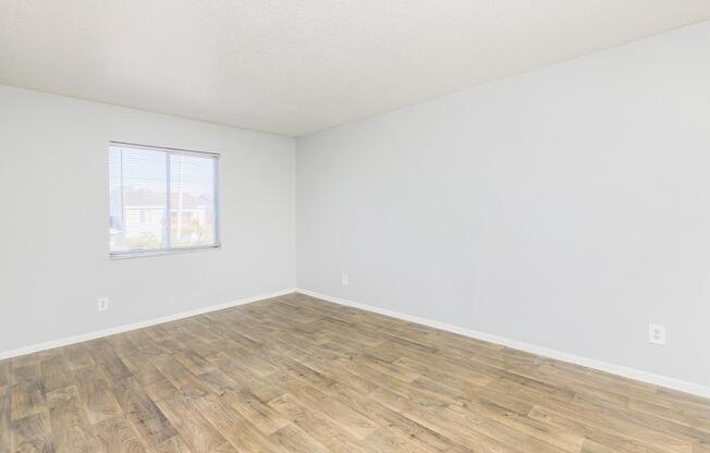 Empty room with light gray walls and a large window with blinds. The flooring is light wood, and the space is well-lit but devoid of furniture or decorations.