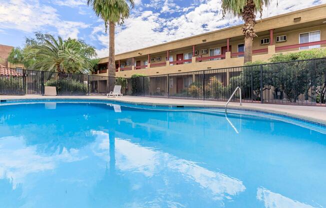 A tranquil swimming pool surrounded by palm trees, with a two-story building in the background. The sky is partly cloudy, reflecting blue tones on the water's surface. A lounge chair is positioned nearby, creating a relaxed atmosphere for leisure and enjoyment.