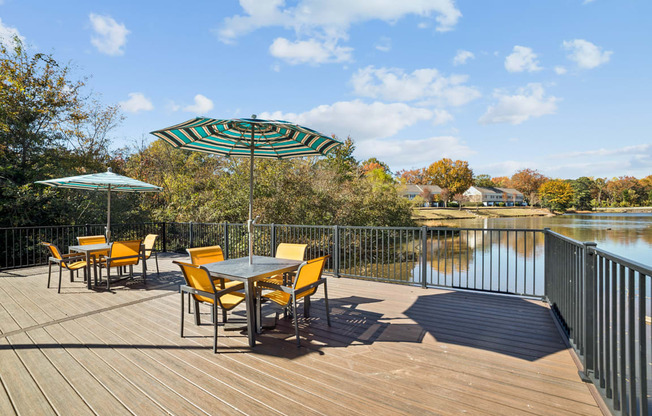 A table with chairs and an umbrella on a wooden deck overlooking a lake.