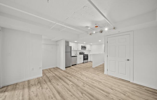 A spacious, modern basement apartment featuring light hardwood flooring, a kitchen with stainless steel appliances, white cabinetry, and bright lighting. The open layout includes a living area and a door leading to another room or closet space. The walls are painted in a crisp, clean white.