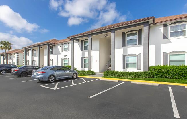A modern apartment complex featuring multiple units with white walls and gray accents. The image shows parking spaces, a well-maintained landscape with green shrubs, and a clear blue sky with scattered clouds. A few parked cars are visible along the front of the building.