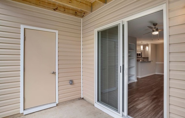 A patio area with concrete floors, tan siding of the exterior building, a door leading to storage, and a sliding glass door leading to the vacant living room with hardwood style flooring.