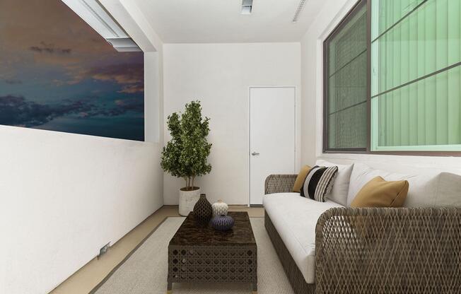 A cozy balcony space featuring a rattan sofa with decorative cushions, a dark coffee table, and a potted plant. In the background, a window shows a cloudy sky at dusk, enhancing the tranquil atmosphere of the area. The walls are painted white, and the flooring is light-colored.