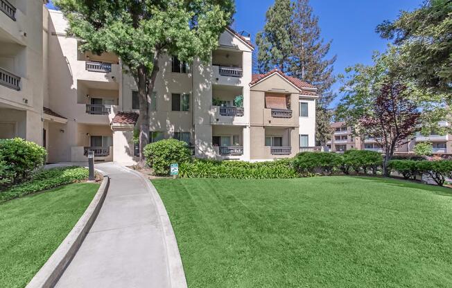 A well-maintained walkway leads through a lush green lawn towards an apartment building surrounded by trees and shrubs. The building features multiple balconies and large windows, with a clear blue sky overhead.