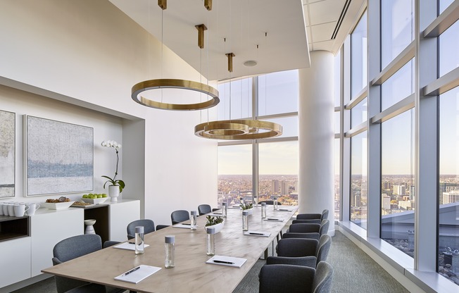 A conference room with a long table and chairs, a kitchen area, and a view of the cityscape through large windows.
