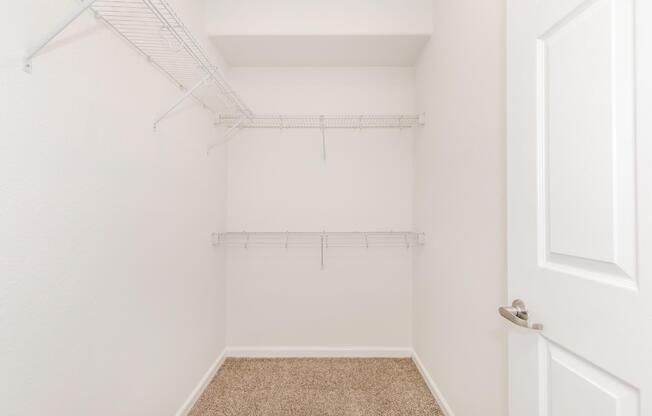 A clean, empty closet with white wire shelving along the walls. The floor is covered in light brown carpet, and there is a white door that is partially closed. The walls are painted a light color, giving the space a bright and airy feel.