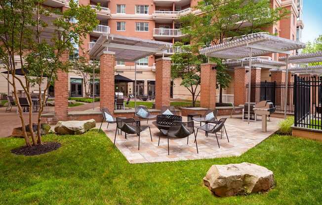 A patio with chairs and a table is surrounded by a grassy area and a building in the background.