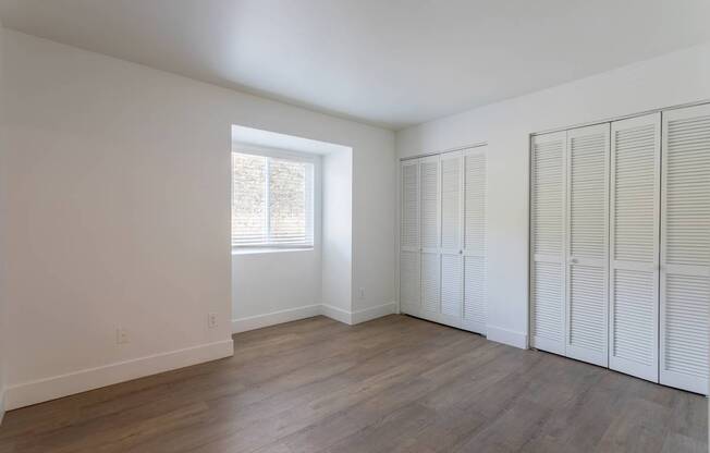 A room with white walls and wooden floors, with a window on the left and a set of white closet doors on the right.