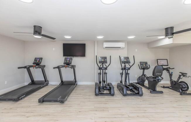 A modern gym featuring three treadmills, two elliptical machines, and a stationary bike, with ceiling fans and wall-mounted TVs in a bright, minimalist space. The flooring is light wood, enhancing the contemporary atmosphere.