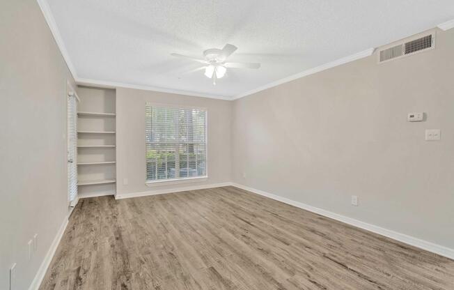 A spacious, empty living room with light-colored walls, a ceiling fan, and laminate flooring. A large window lets in natural light, and there are built-in shelves on one side. The room is well-lit and invites a welcoming atmosphere.