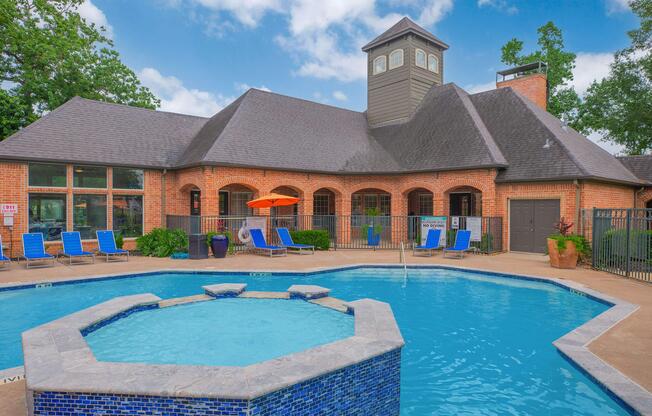 A well-maintained swimming pool surrounded by lounge chairs, featuring a hot tub in the foreground. In the background, there is a large brick building with a peaked roof and large windows, shaded by trees. An orange umbrella adds a pop of color to the outdoor area. Bright blue sky enhances the inviting atmosphere.