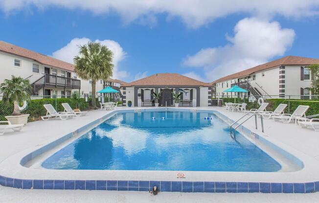 A crystal-clear swimming pool surrounded by lounge chairs and palm trees, with two buildings featuring green umbrellas in the background. The sky is partly cloudy, creating a serene and inviting atmosphere.