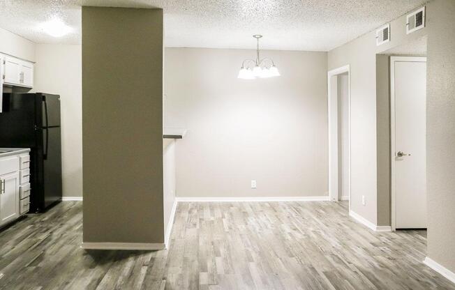 A well-lit, modern kitchen and living area featuring light wood flooring. The kitchen has white cabinets and a black refrigerator. A dining area is indicated with a hanging light fixture above. A door leads to another room, and the walls are painted a neutral color, creating an inviting atmosphere.