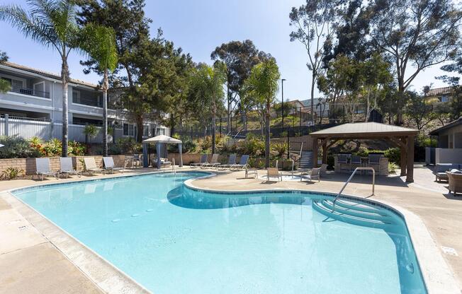 Outdoor swimming pool surrounded by palm trees and lounge chairs. A shaded gazebo is nearby, along with landscaping featuring plants and bushes. The area is sunny and well-maintained, with a residential building visible in the background.