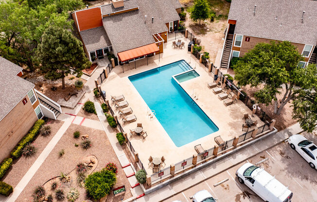 Aerial view of the outdoor pool area here at Trinity Place with a rectangular swimming pool centered within a fenced courtyard. Lounge chairs and patio tables surround the pool deck, while nearby apartment buildings, shaded walkways, and mature trees frame the community space and create a relaxed, resort-style setting.