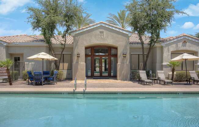 A pool in front of a house with a patio and chairs.