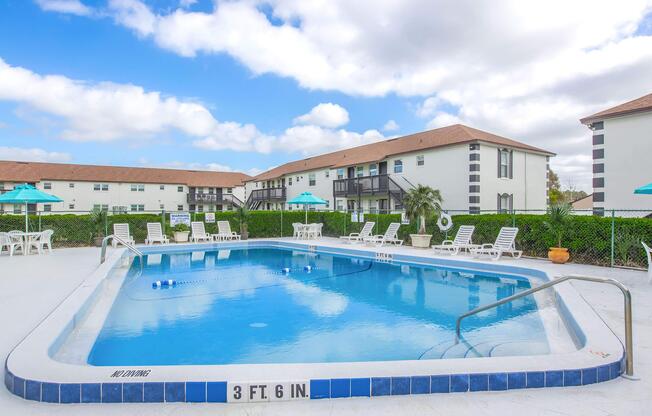 A clear blue swimming pool surrounded by white lounge chairs and umbrellas. In the background, there are two-story apartment buildings with a light color scheme. The sky is partly cloudy, creating a bright and inviting atmosphere. A depth marker shows the pool is 3 feet 6 inches deep.