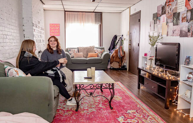 two women sit in a living room with a red rug and a television