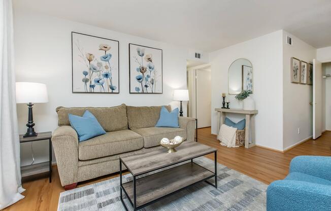 Cozy living room featuring a beige sofa with blue cushions, a wooden coffee table, and soft lighting from table lamps. Beautiful floral artwork adorns the white walls, and a minimalist console table with decorative items is present. The room has warm wood flooring and neutral tones for a welcoming feel.
