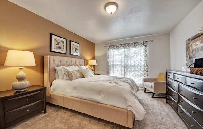 A welcoming bedroom here at High Plains with plush beige carpet, a neutral upholstered bed, warm accent walls, and soft lighting, complemented by a large window with patterned curtains and tasteful furnishings that create a calm, comfortable retreat.