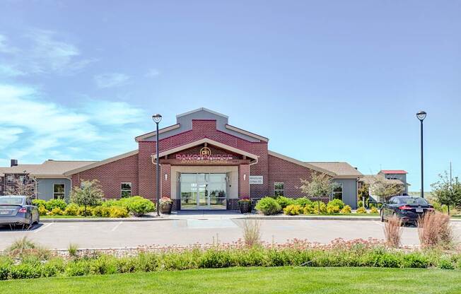 A welcoming exterior view here at Dakota Ridge featuring the brick clubhouse entrance with a peaked roofline, glass double doors, landscaped greenery, colorful planters, and a wide parking area set against a bright blue sky and well-maintained lawns.