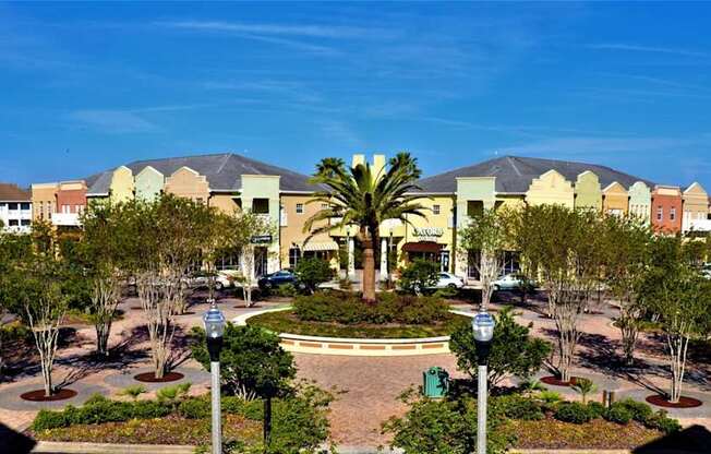A large building with a yellowish hue is surrounded by trees and a fountain in the middle of a plaza.
