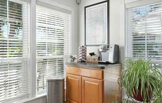 A room with a window covered in blinds, a plant, a framed map, and a microwave on a counter.