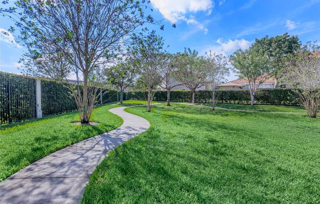 A winding concrete pathway leads through a lush green lawn, bordered by small trees with sparse foliage. The background features a tall hedge and a clear blue sky with a few clouds, creating a serene outdoor setting ideal for relaxation or a leisurely stroll.
