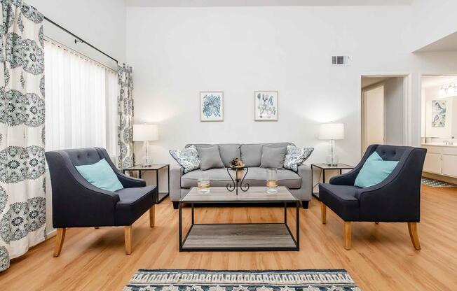 A modern living room featuring a gray sofa with decorative pillows, two stylish black armchairs with teal accents, a glass coffee table with decorative items, and light-colored curtains. The walls are adorned with framed artwork, and there are lamps providing soft lighting in the space.