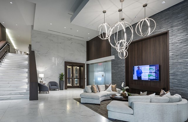 A grand lobby space featuring high ceilings, marble floors and walls, a marble staircase, a modern chandelier of interlocking rings, and a seating area with wood-paneled walls, an HDTV, and two large, white, curved couches.