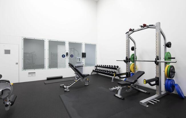 A modern gym interior featuring a weight bench with a barbell, dumbbells arranged on a rack, and a multi-gym machine. The flooring is black rubber, and there are large glass mirrors on one wall. The overall design is minimalist and functional, suitable for strength training.