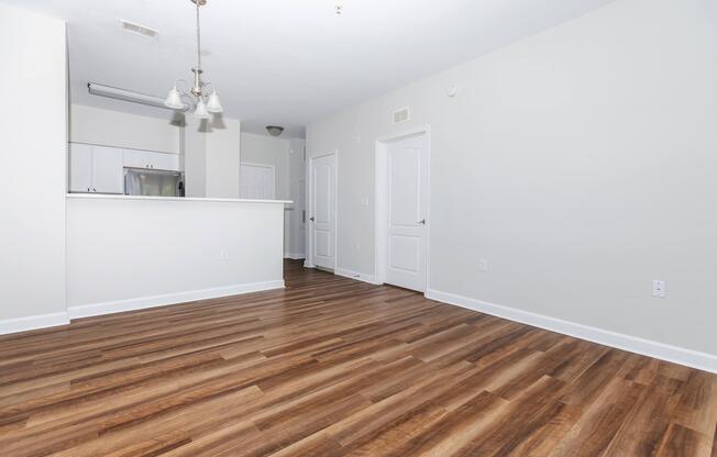 A spacious, empty living area with light wood flooring, white walls, and a kitchen area in the background. There are modern light fixtures hanging from the ceiling, and two doorways lead to other rooms. Natural light flows in, creating a bright atmosphere.