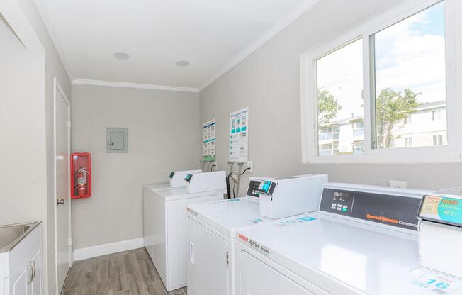 A laundry room featuring several white washing machines and dryers, with a window providing natural light. The walls are painted a light gray, and there is a fire extinguisher mounted on the wall. Various informational posters are displayed near the machines.