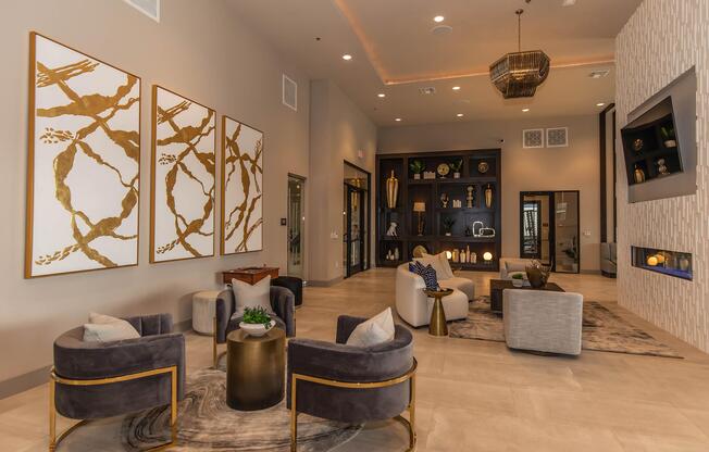 Modern lounge area featuring elegant decor with two large gold and white abstract wall art pieces. Stylish gray chairs are arranged around a round gold table, with a lush plant centerpiece. A cozy fireplace and built-in shelving with decorative items enhance the inviting atmosphere.
