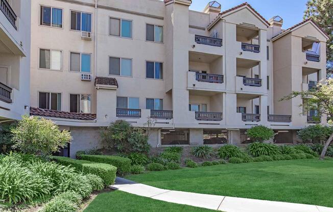 A residential building with multiple floors featuring balconies. The exterior is light-colored with a tiled roof. There is a well-maintained green lawn in the foreground with lush plants and shrubs. Windows are visible on each floor, and some balconies have potted plants. Sunlight brightens the scene.