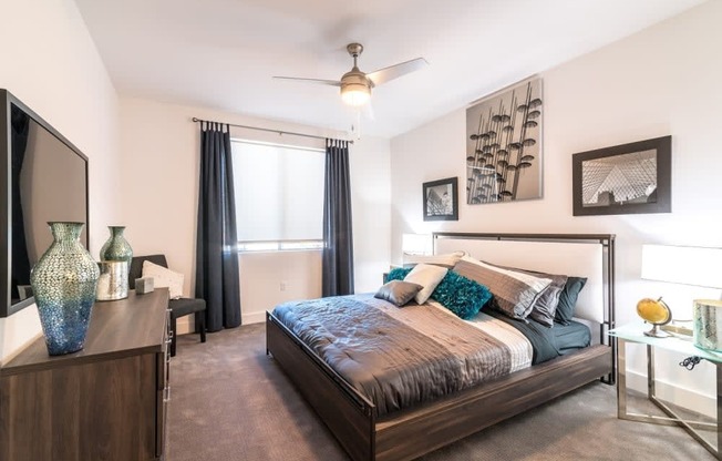 A bedroom with a tv and dresser and window and ceiling fan and large bed and carpet