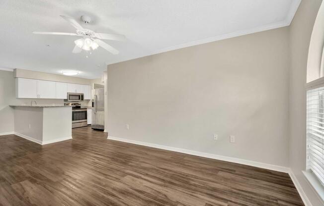 Spacious living area with hardwood floors and light-colored walls. A ceiling fan is visible, and the room opens to a modern kitchen featuring stainless steel appliances. There are large windows allowing natural light, creating a bright and inviting atmosphere.