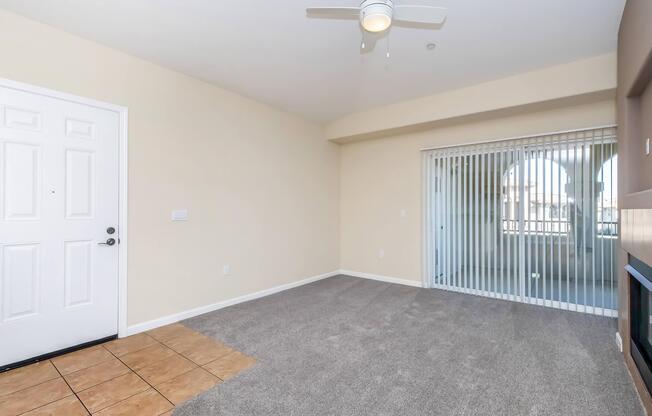 A bright, vacant interior space featuring light-colored walls, a ceiling fan, and a sliding glass door with vertical blinds leading to a balcony. The floor includes beige tiles and a carpeted area, creating a cozy living room atmosphere.