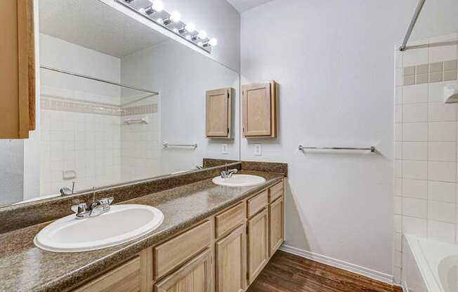Bright and spacious bathroom at Saxony at Chase Oaks Apartments in Dallas, TX, featuring dual sinks, a large mirror, and wood-style flooring.