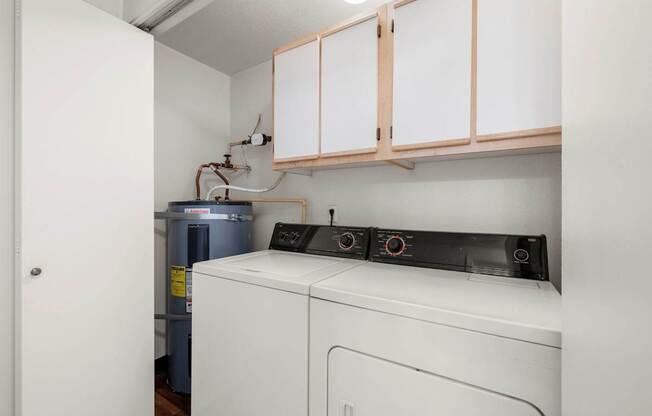 A white washing machine sits under a cabinet in a small room at Clackamas Trails Apartments, Oregon, 97222
