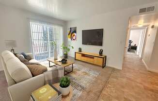 Bridgeside Landing Apartments in Taylorsville, Utah, Living area with tan couch, flatscreen television on wall, balcony off the room, hallway back to bedroom.