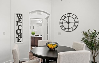 A dining room with a black table and chairs, a clock on the wall, and a "Welcome" sign on the door.