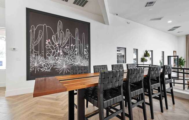 A long wooden table is surrounded by chairs in a room with a desert painting on the wall.