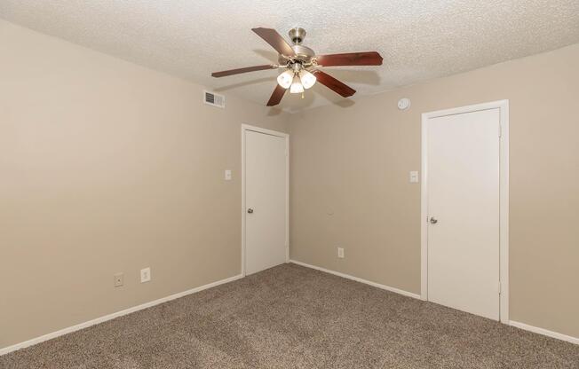 A neutral-colored bedroom featuring a textured ceiling with a ceiling fan, plush carpeted flooring, and two closed white doors on adjacent walls. The room has minimal decor, creating a spacious and simple atmosphere.